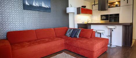 Una sala de estar moderna con un sofá seccional rojo, un muro con diseño y una zona de cocina con electrodomésticos blancos.