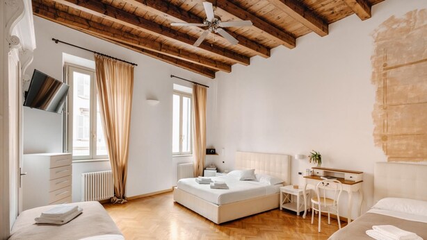 Une chambre avec un grand lit, un bureau et une chaise. Il y a une fenêtre avec des rideaux, un radiateur et un ventilateur de plafond.