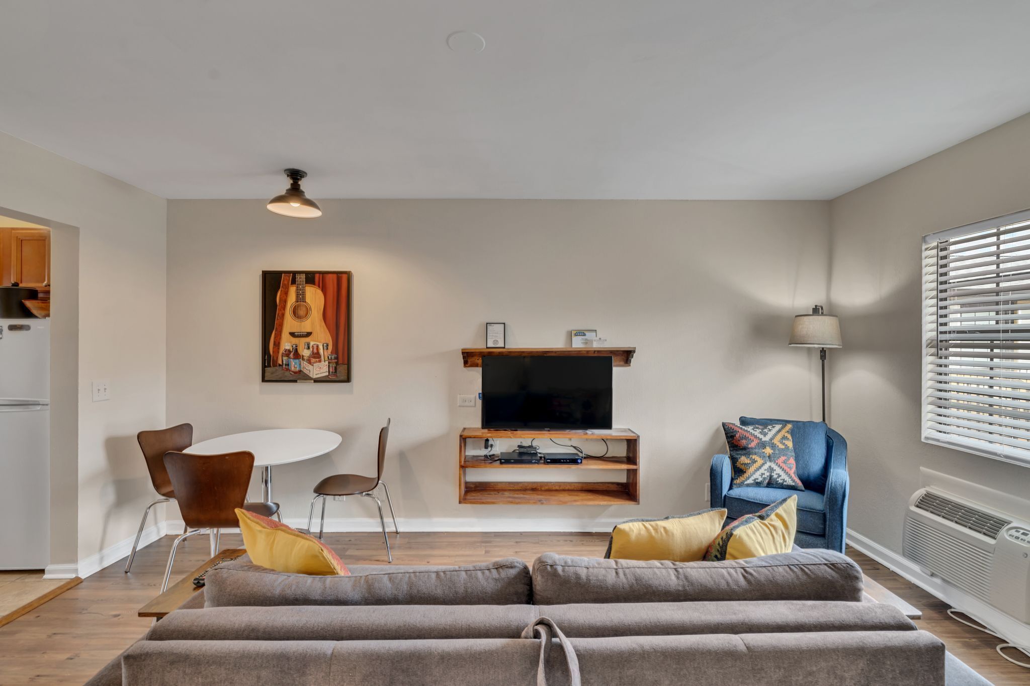 A modern living room with a grey sofa, a dining table with chairs, a TV on a wooden stand, and a window with blinds.