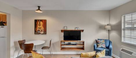 A modern living room with a grey sofa, a dining table with chairs, a TV on a wooden stand, and a window with blinds.