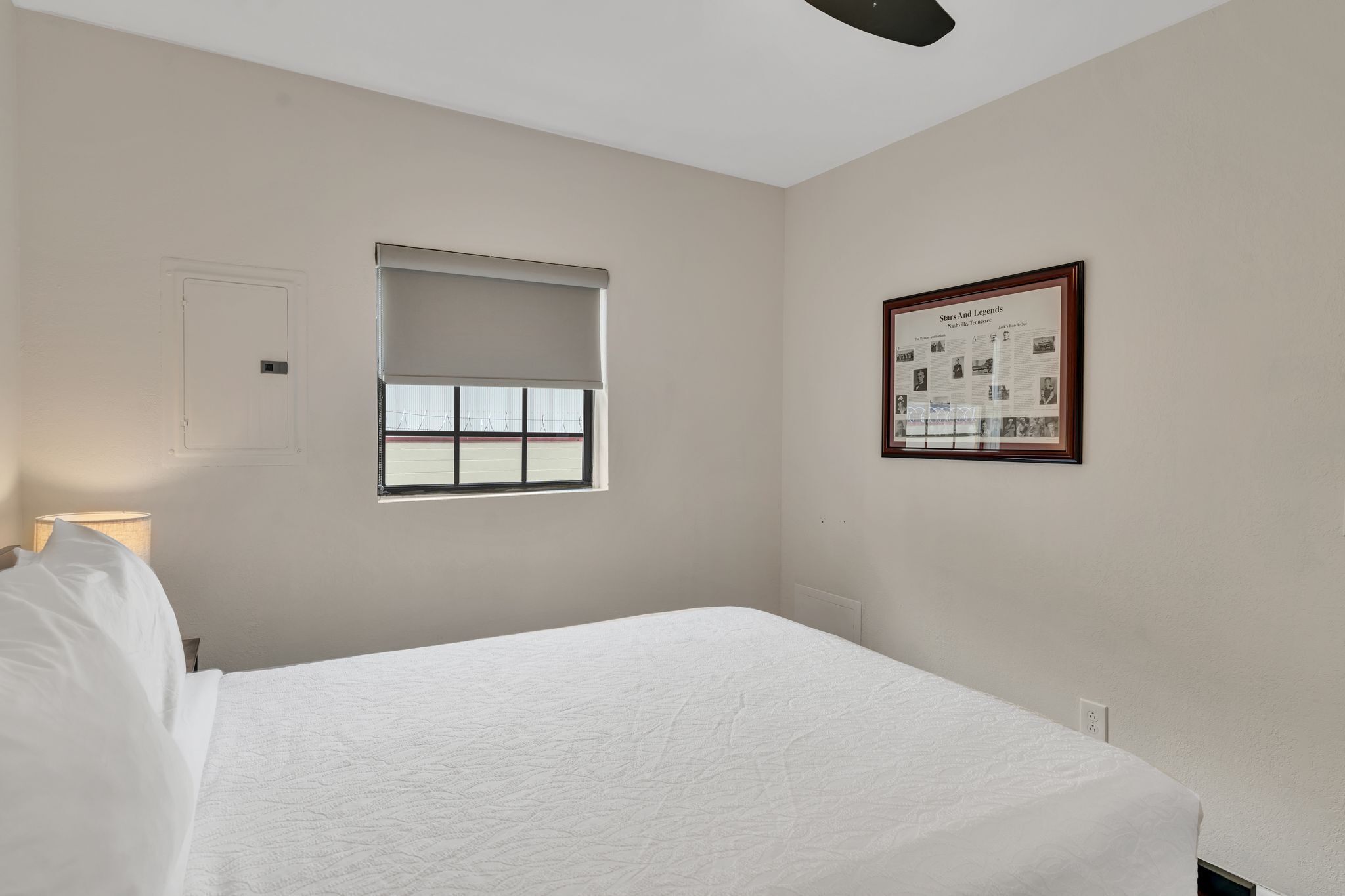 A bedroom with a bed, a window with blinds, a wall-mounted light, and a framed picture.