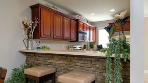 A kitchen with wooden cabinets, a stone countertop, two stools, and a hanging plant. - RiverHaus on Edelweiss - Cozy River Condo Downtown (Helen)