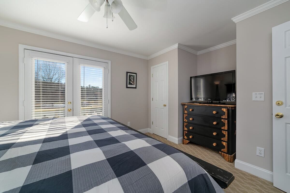 A bedroom with a checkered bedspread, a ceiling fan, a dresser with drawers, a television, and a door with a window.