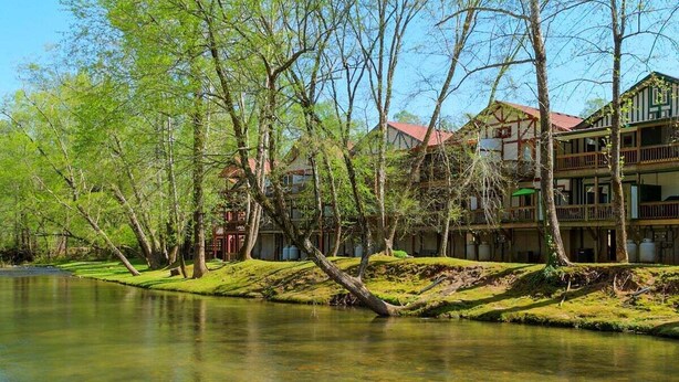 A riverside hotel with multiple floors, a grassy bank, and trees. - RiverHaus on Edelweiss - Cozy River Condo Downtown (Helen)