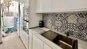 A modern kitchen with white cabinets, a patterned backsplash, a stainless steel sink, and a view of palm trees outside.