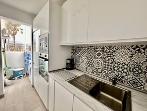 A modern kitchen with white cabinets, a patterned backsplash, a stainless steel sink, and a view of palm trees outside.