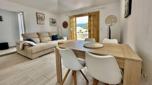 Een moderne woonkamer met een houten eettafel, witte stoelen, een beige bank en uitzicht door een open deur.