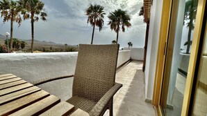 Een balkon met een rieten stoel en een houten tafel, met uitzicht op palmbomen en een uitgestrekt landschap.