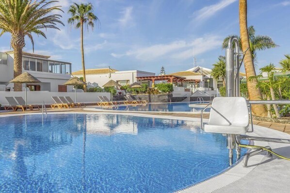 Une piscine avec des chaises longues, des palmiers et un bâtiment doté d’un toit en tuiles.