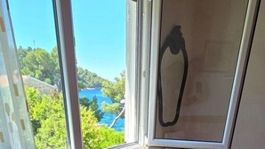 A bedroom with a wooden floor, a bed with a pink and white floral bedspread, a window with a view of greenery and a blue structure, and a chair with a white cover. - Apartment Coraline on Korcula, seafront (Smokvica)