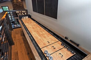 Una mesa de juego de shuffleboard con zonas de puntuación numeradas, un suelo de madera y una pared de piedra de fondo.