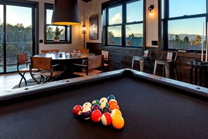 Una mesa de billar con bolas, un comedor con sillas y una mesa, y amplias ventanas con vistas al exterior.
