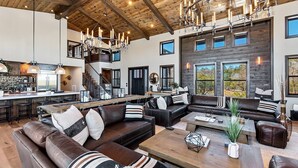 A spacious living area with a high ceiling, a central seating area with sofas and a coffee table, a kitchen area with a bar, and large windows offering views of the outdoors.