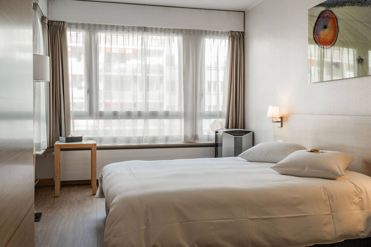 A hotel room with a bed, a bedside table, a lamp, and a window with curtains.