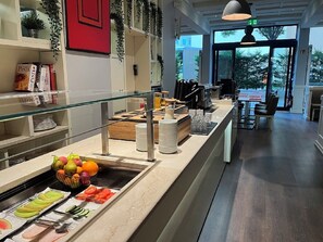 Een buffetgedeelte met diverse soorten fruit en groenten, een pastasalad en een zithoek met tafels en stoelen.
