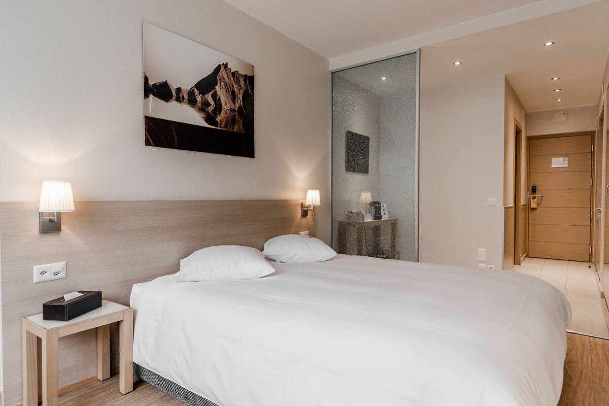 A modern hotel room with a large bed, bedside lamps, a nightstand, a wall-mounted painting, and a mirror.