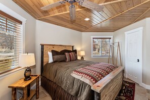 Un dormitorio con techo de madera, cama con ropa de cama estampada, mesita de noche con lámpara y ventana con persianas.