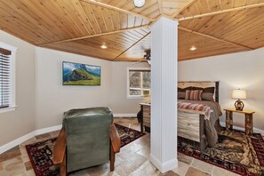 A bedroom with a wooden ceiling, a bed with a patterned bedspread, a green leather chair, and a painting of a landscape.