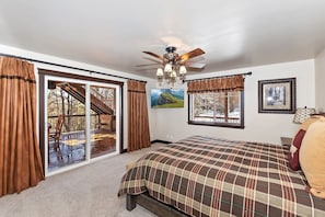 A bedroom with a large bed, a ceiling fan, a window with curtains, and a view of a deck.