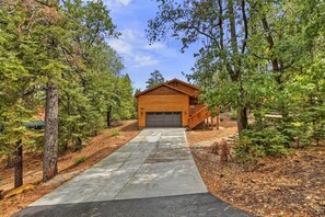 A log cabin with a garage, surrounded by dense trees and a concrete pathway leading to it.