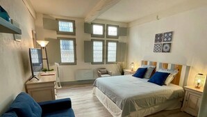A bedroom with a large bed, a blue sofa, a desk with a lamp, and multiple windows. - 'The Ucetia' appt in charming stone house in Uzès centre (Uzès)