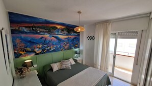 Une chambre avec une grande fresque murale représentant un coucher de soleil sur l’eau, un lit avec une tête de lit verte, une table de chevet avec une lampe, et une vue sur l’extérieur grâce à une porte coulissante.