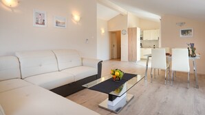Un salon moderne avec un canapé blanc, une table basse en verre sur laquelle sont disposés des fruits, et un coin repas avec des chaises blanches.