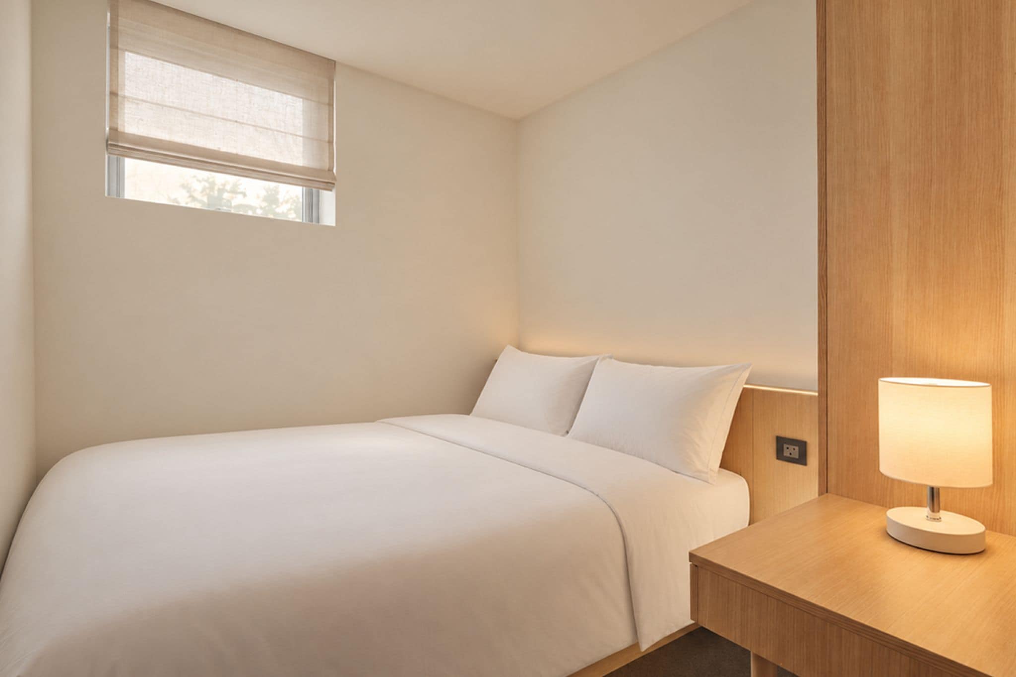 A hotel room with a bed, a bedside table with a lamp, and a window with blinds.