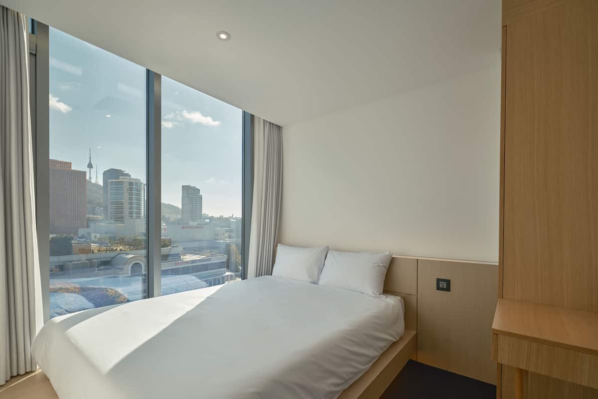 A hotel room with a large bed, a wooden nightstand, and a view of a cityscape through large windows.