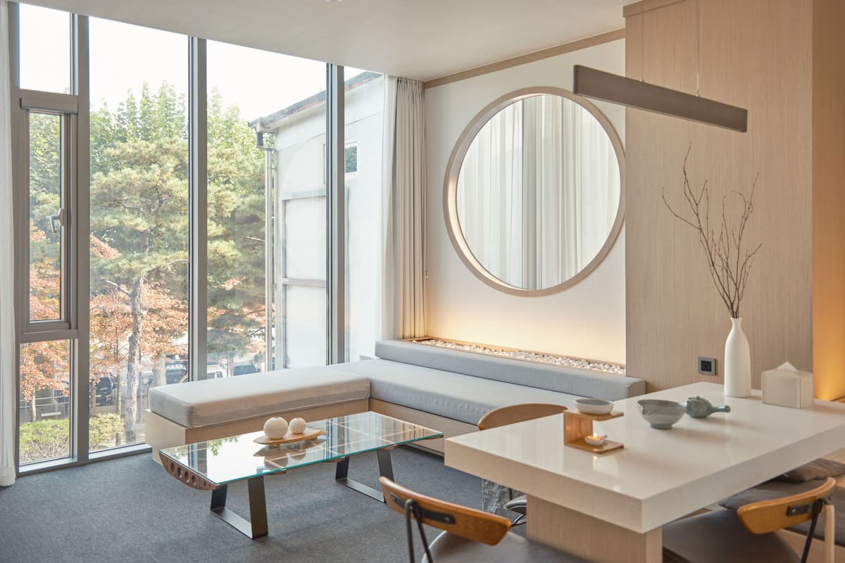 A modern dining area with a glass table, cushioned seating, and a view of trees through large windows.