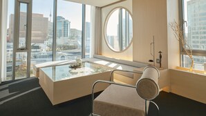 A modern hotel room with a large window, a round mirror, a glass table, and a cushioned chair. - UH Suite The Seoul (Seoul)