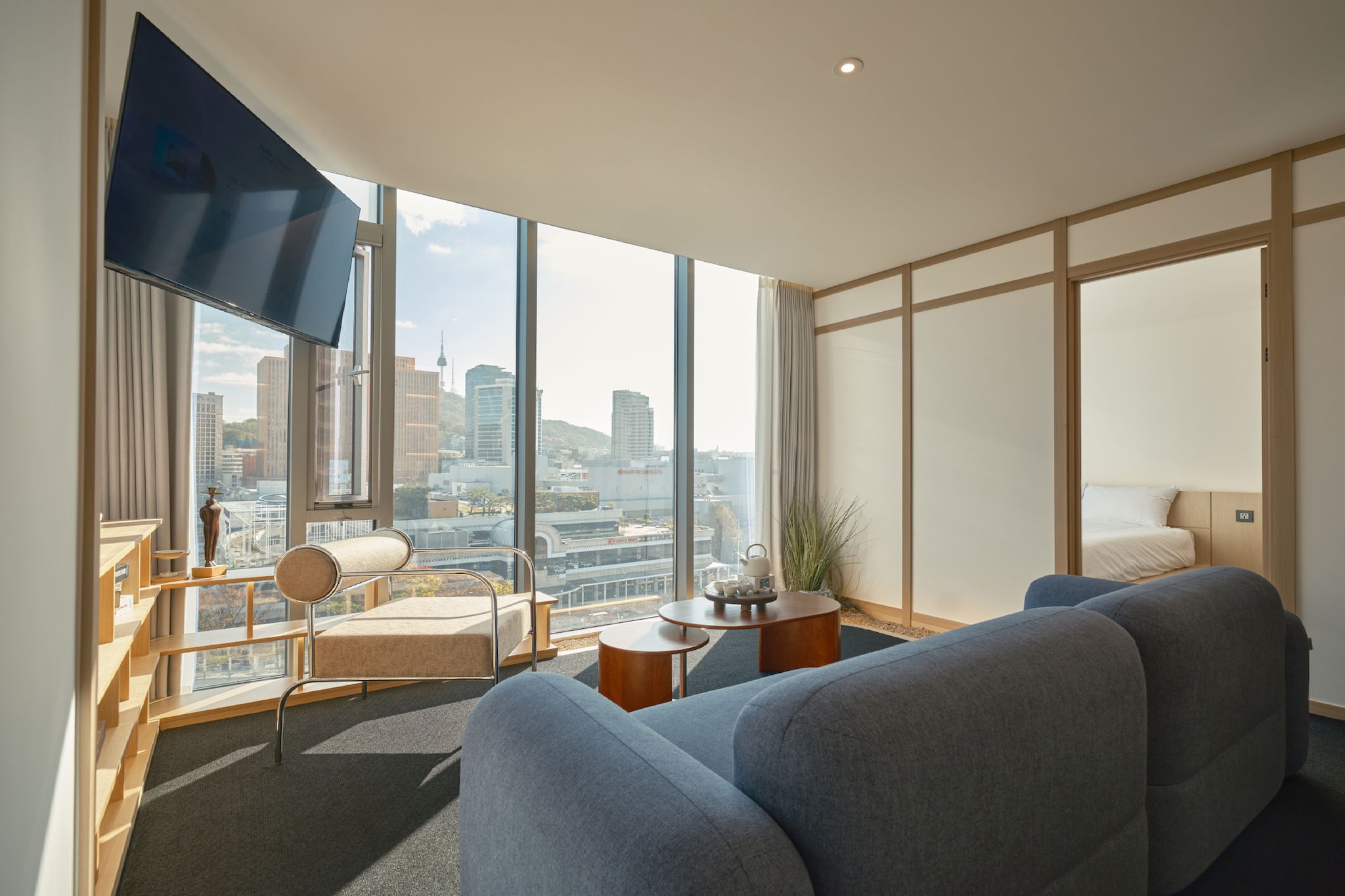 A modern hotel room with a large window, a sofa, a chair, a small table, and a flat-screen TV.