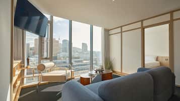 A modern hotel room with a large window, a sofa, a chair, a small table, and a flat-screen TV.