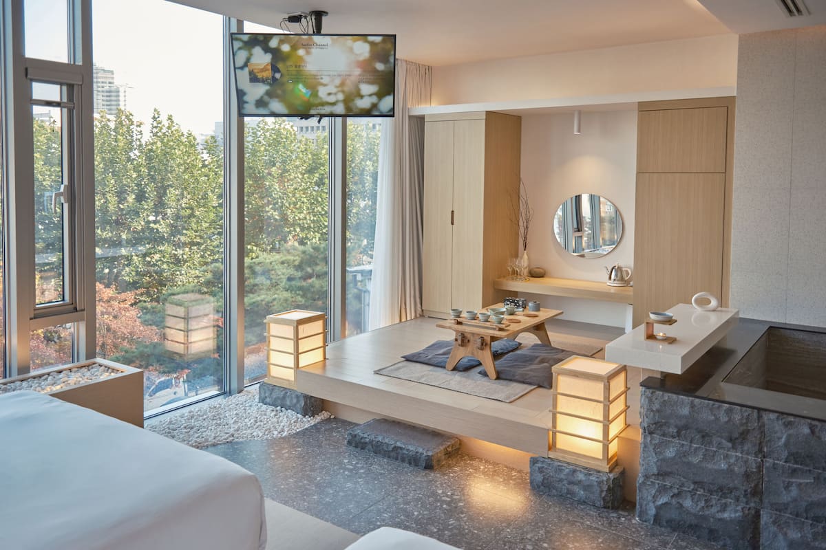 A modern hotel room with a large window, a wooden table with bowls, a mirror, and a stone accent wall.