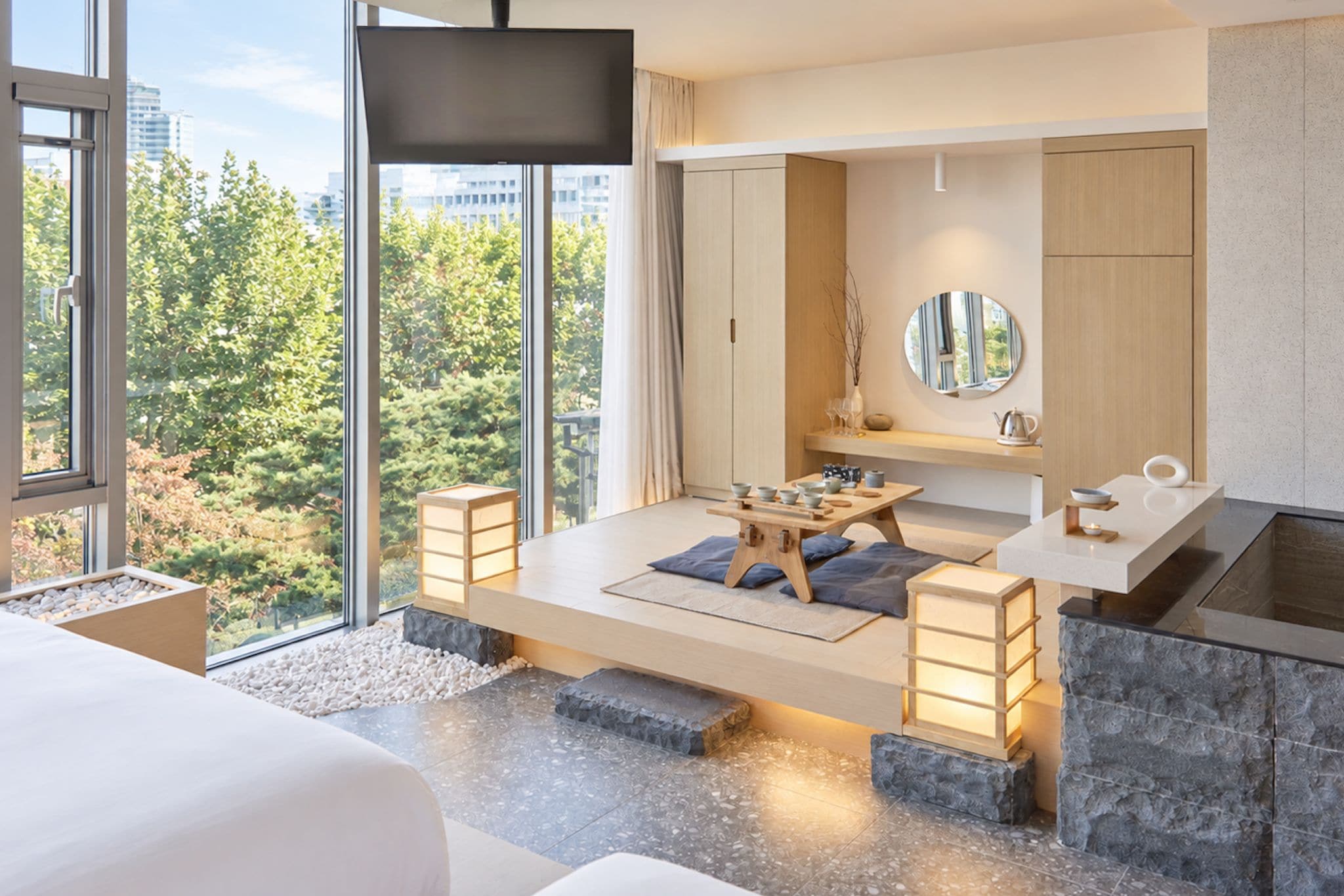 A modern hotel room with a large bed, a dining area, a bathroom, and a view of the city.