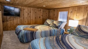 Una habitación con paredes de madera, dos camas con ropa de cama a rayas, un televisor montado en la pared y una ventana con persianas.