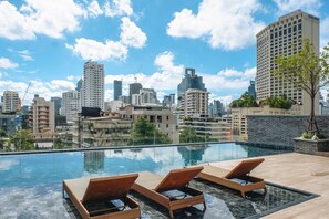 Una piscina infinita en la azotea con sillones de descanso, con vistas a un paisaje urbano de edificios altos.