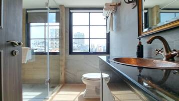 A modern bathroom with a glass shower enclosure, a large window with a city view, and a copper-finished sink.
