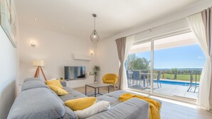 A modern living room with a grey sectional sofa, a flat-screen TV, a small table, and a yellow armchair. A large window offers a view of a swimming pool and greenery. - Villa Defiance (Benkovac)