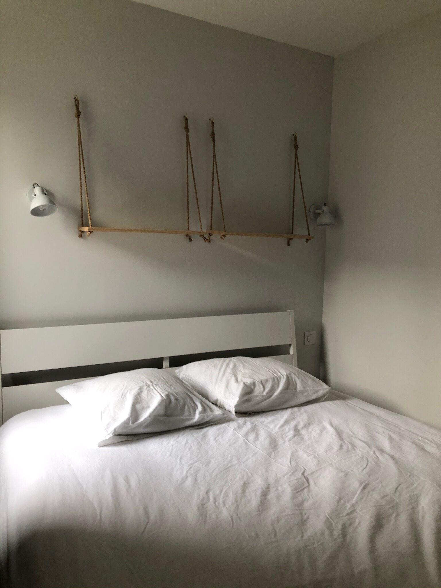 Una cama con ropa de cama blanca y dos almohadas, cabecera blanca y dos lámparas de pared con estructura de madera.