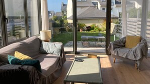 Un salon moderne avec un canapé, un fauteuil et une petite table. De grandes fenêtres offrent une vue sur un jardin et les maisons voisines.