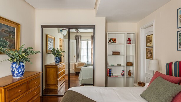 Un dormitorio con cama, cómoda, espejo, estantería y puerta que da a otra habitación.