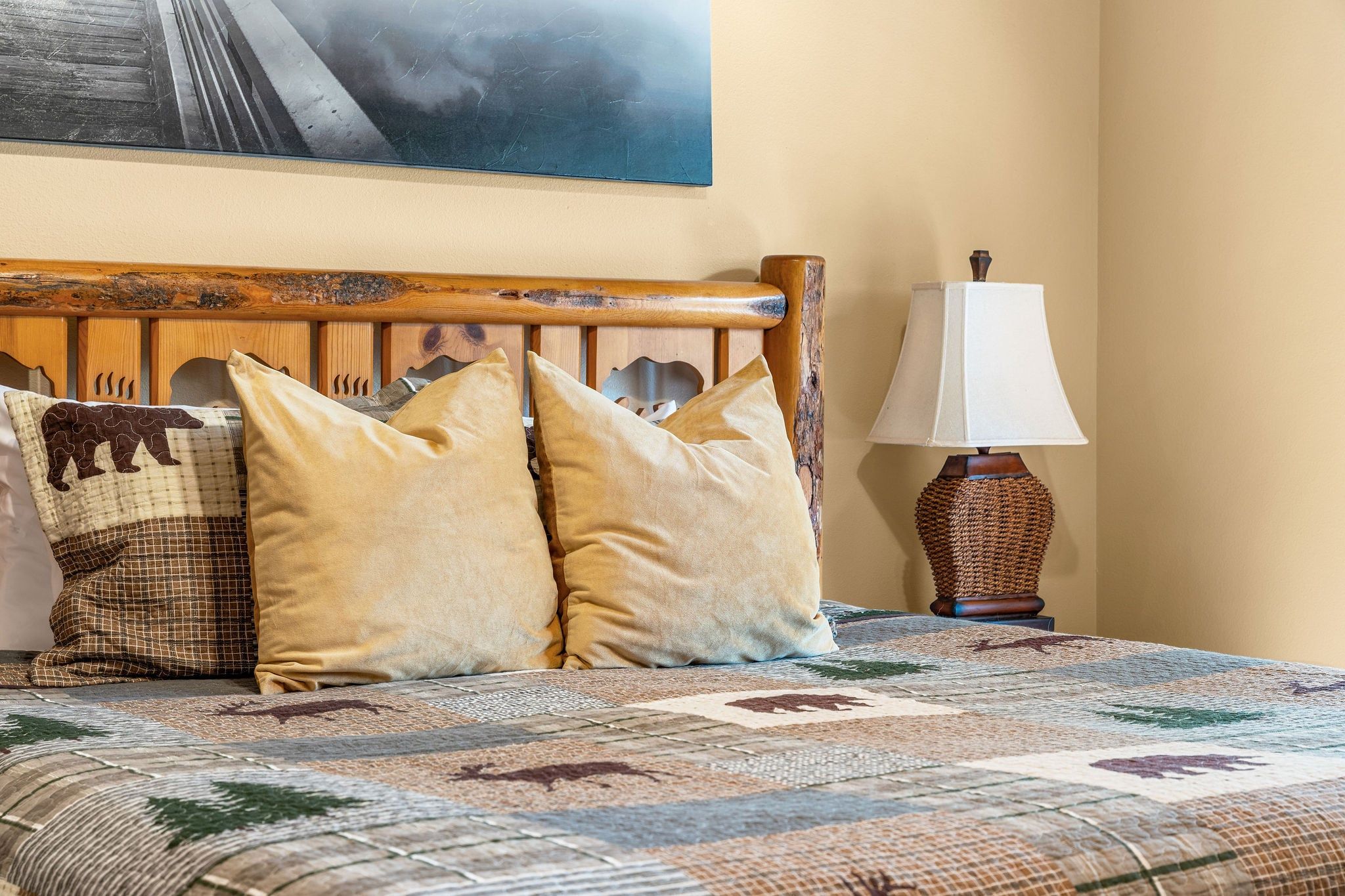 A bed with a patterned bedspread, two pillows, a decorative headboard, a wall-mounted lamp, and a painting above the bed.