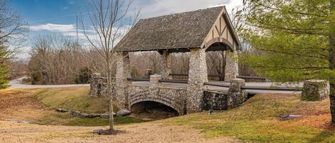 A stone bridge with a covered walkway, surrounded by trees and a grassy area.