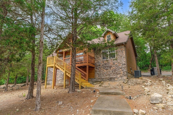 A cabin with a stone exterior, wooden deck, and a staircase leading to a second-story balcony.