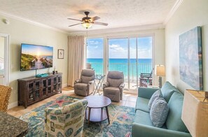A living room with a TV, two recliners, a sofa, a coffee table, and a view of the ocean through large windows.