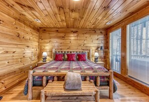 A log cabin bedroom with a wooden bed, pillows, a bench, and a nightstand with a lamp.
