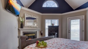 A bedroom with a fireplace, a bed with floral bedding, a large mirror, and a TV on the wall. - Ober Hideaway, Hot Tub, WIFI, $560 FREE tickets each paid day!!! (Gatlinburg)