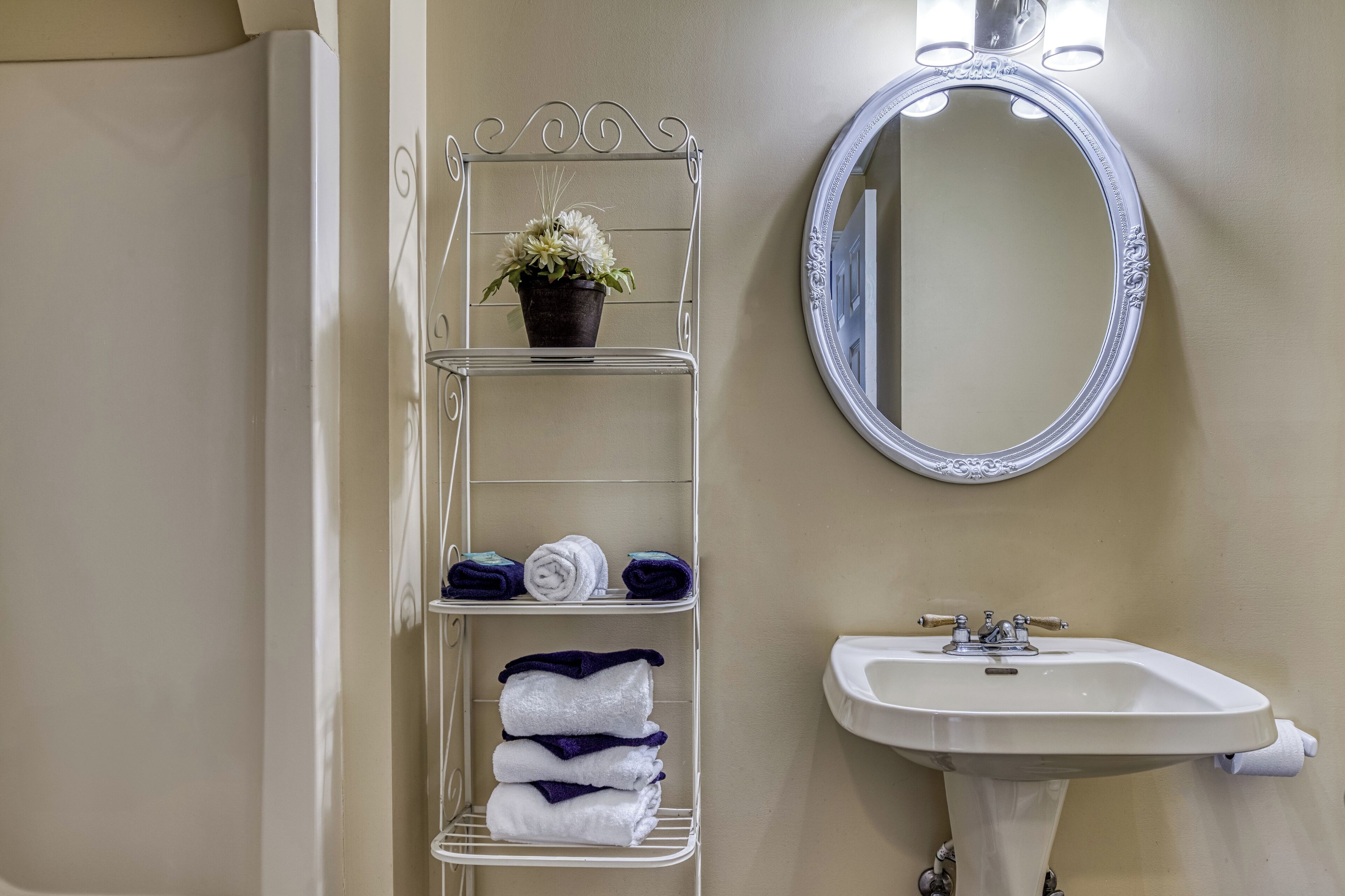 A bathroom with a white metal shelf holding towels and a potted plant, a round mirror, and a white sink.