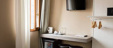 A hotel room with a wooden desk, a flat-screen TV mounted on the wall, a small refrigerator, and a trash bin.